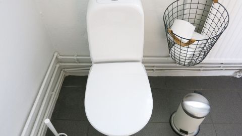 Schmunzeltipp, zum Toilettenwasser sparen: Mit einem Backstein im Spülkasten Wasser beim Spülen einsparen. Ein modernes, weißes WC steht in einem schlicht gestalteten Raum, daneben befinden sich eine Abfallkammer und eine Toilettenbürste.