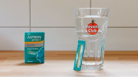 Vermeide Katerstimmung, indem du vor dem Schlafen ein Glas Wasser und ein Aspirin nimmst. Dies hilft, den Flüssigkeitsverlust auszugleichen und Kopfschmerzen vorzubeugen. Ein einfacher Trick für einen klaren Morgen. Eine Aspirin-Effect-Packung steht links neben einem Havana Club Glas mit Wasser, passend zum Thema Katerstimmung vermeiden.
