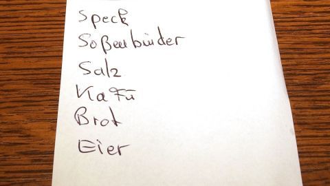 Mit Einkaufsliste und anderen Tipps beim Einkaufen sparen. Eine handgeschriebene Einkaufsliste auf einem Notizblatt zeigt verschiedene Lebensmittel wie Speck, Salz und Eier zur Planung von Sparen beim Einkauf.