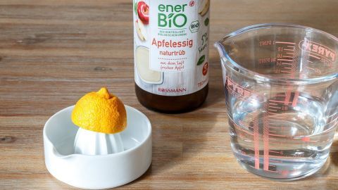 Mithilfe von Apfelessig und Zitronensaft einen guten und günstigen Haarfestiger herstellen. Ein Fläschchen Apfelessig steht neben einem Eierbecher mit einem halben Zitrusfruit und einem Messbecher mit Wasser.