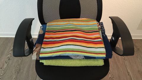 Bettwäsche bügeln ganz einfach während ich arbeite. Einfach die Bettwäsche zwischen zwei Handtücher legen und drauf sitzen. Ein Stapel mit bunt gestreiften Handtüchern liegt ordentlich auf einem schwarzen Bürostuhl in einem hellen Raum.