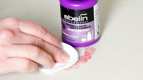Mit acetonfreiem Nagellack kannst du Make-up-Flecken aus weißem Kunstleder entfernen. Eine Hand reinigt einen roten Fleck von weißem Kunstleder mit acetonfreiem Nagellackentferner.