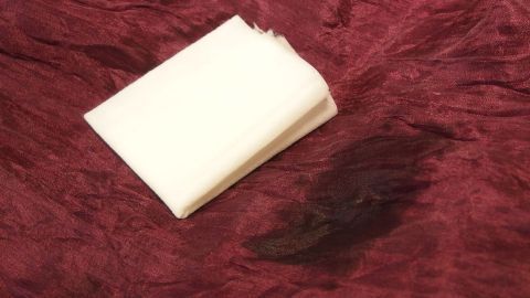 Frische Schokoladenflecken, Dreckflecken, Soße, etc. lassen sich prima mit Feuchttüchern entfernen. Ein Feuchttuch liegt auf einem roten Stoff mit einem frischen Fleck daneben, um eine mögliche Reinigung zu zeigen.