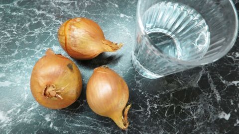 Zwiebeln schneiden ohne Weinen? Einfach einen Schluck Wasser in den Mund nehmen und dann ganz normal die Zwiebel schneiden. Zwiebeln liegen auf einer dunklen Marmorfläche neben einem klaren Wasserglas, bereit zum Schneiden und Kochen.