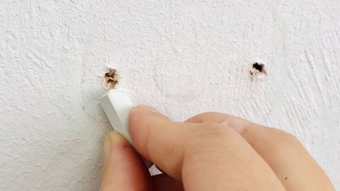 Kleine Löcher in der Wand von Reißzwecken oder Nägeln verschwinden ganz schnell mit etwas weißer Kreide. Eine Hand hält ein Stück Kreide und „versiegelt“ kleine Löcher in einer weißen Wand. Der Hintergrund ist schlicht und hell.
