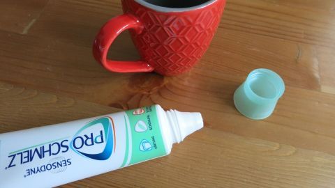 Ein einfacher Haushaltstrick: Mit Zahnpasta lassen sich getrocknete Kaffeeränder entfernen. Trage etwas Zahnpasta auf den Fleck auf, reibe sanft und wische es dann ab. So bleibt dein Tisch sauber und frisch. Zahnpasta liegt offen neben einer roten Tasse auf einem Holztisch. Text auf der Tube: SENSODYNE PRO SCHMELZ.