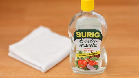 Tränke ein Tuch mit Essigwasser, lege es auf die glänzende Stelle und bügle darüber. Der Stoff ist sofort wieder matt. Eine Flasche Essig und ein Tuch liegen auf einem Holztisch, bereit zum Entfernen von Glanzstellen vom Bügeln.