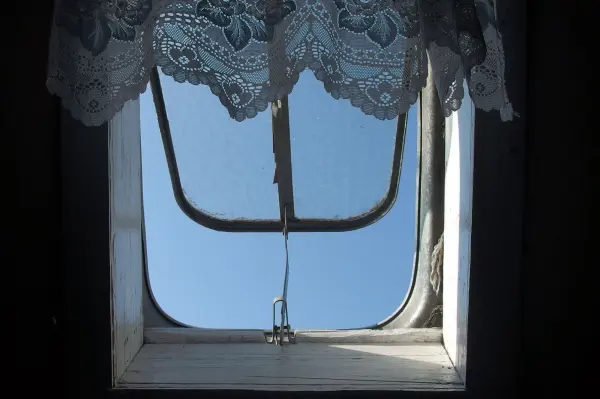 Ein Dachfenster im geöffneten Zustand zeigt den blauen Himmel, umgeben von einer Spitzenvorhang. Es wirkt schmutzig und benötigt Reinigung.
