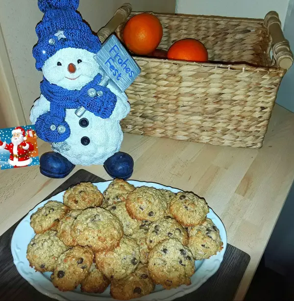 Ein Teller Haferflocken-Cookies steht auf einem Tisch. Daneben ein Schneemann mit Schild "Frohes Fest", vor einem Korb mit Orangen.
