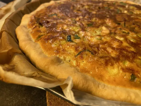 Eine appetitliche, saftige Gemüse-Quiche liegt frisch gebacken in einer Form auf einem Tisch, umgeben von Backpapier.