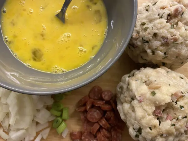 Geröstete Knödel liegen neben einer Schüssel mit aufgeschlagenen Eiern, umgeben von Zwiebeln und Wurststücken.