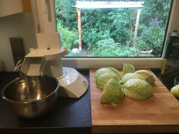 Auf einem Küchenarbeitsplatz liegen geschnittene Weißkohlblätter, während eine Küchemaschine zum Zubereiten von Krautsalat bereitsteht.