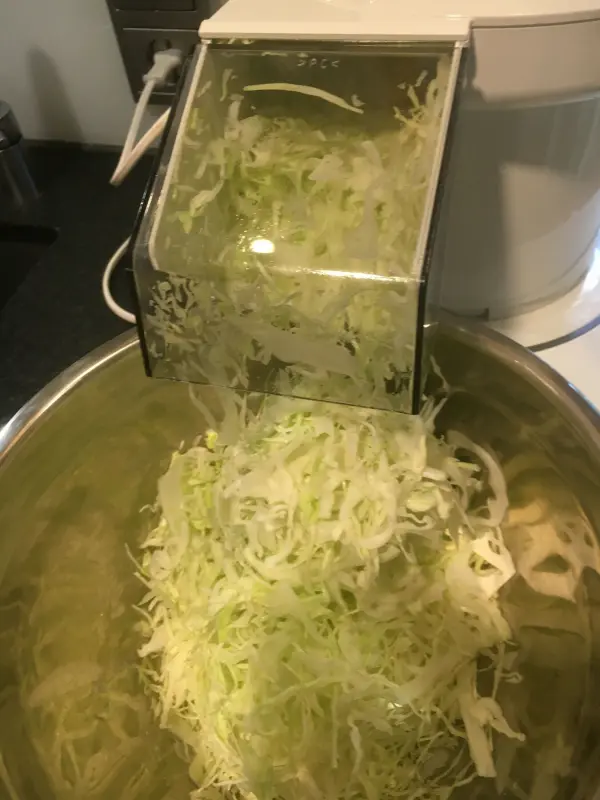 Geriebener Weißkohl fällt in eine große Schüssel, während ein Küchenhelfer das Gemüse zubereitet.