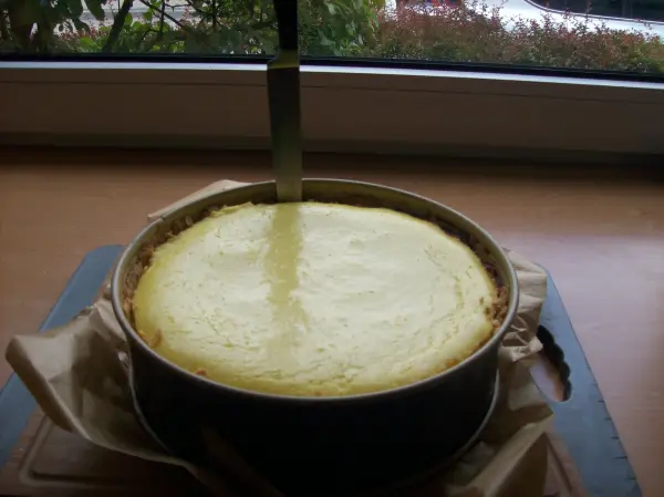 Ein Käsekuchen steht auf einem Tisch und wird gerade mit einem Kuchenschneider angeschnitten, um ihn portionsweise zu servieren.
