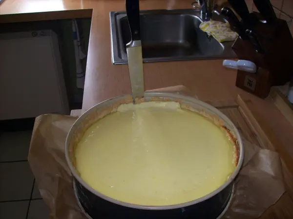 Ein Käsekuchen steht in einer Küche auf einer Arbeitsfläche. Ein Messer schneidet ihn vorsichtig an, während im Hintergrund Geschirr und eine Spüle sichtbar sind.