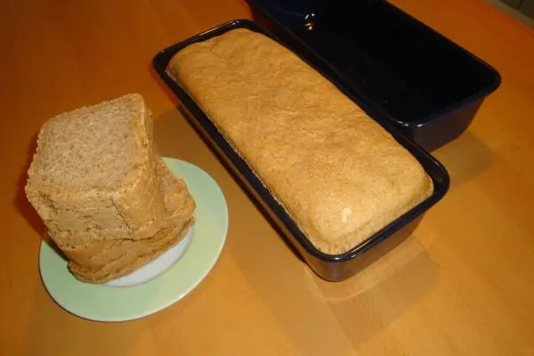 Ein dunkles Dinkelbrot liegt in einer Kastenform, während mehrere Scheiben auf einem Teller daneben gestapelt sind.