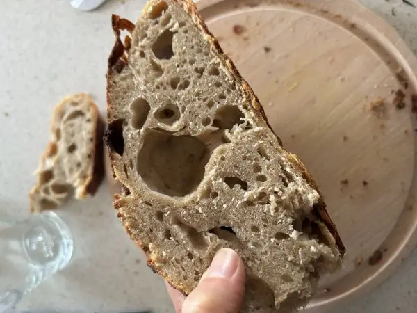 Ein Stück Dinkel-Sauerteigbrot wird auf einer Küchenplatte gehalten, daneben ein rundes Holzbrett mit Brotkrümeln.