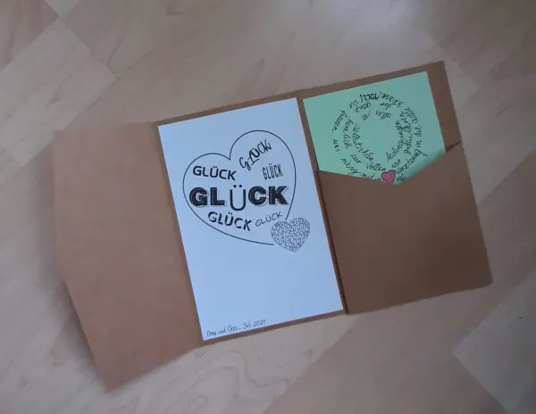 Eine Glückwunschkarte liegt geöffnet auf einem Tisch, mit einem herzförmigen Design, das "Glück" betont, und einem kleinen, beschriebenen Zettel in einer Tasche.