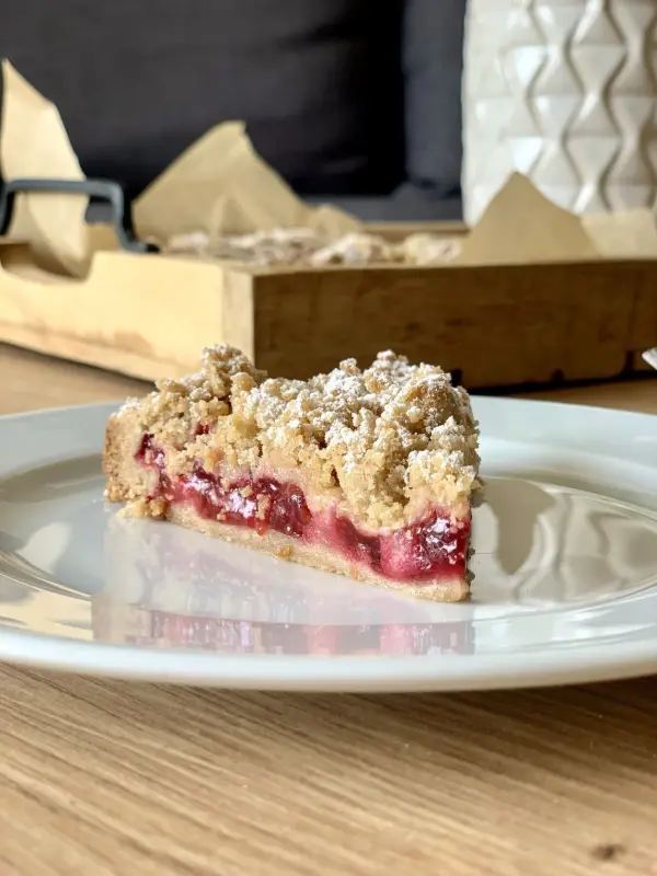 Ein Stück Streuselkuchen liegt auf einem weißen Teller, umgeben von Holz und sanften, grauen Akzenten im Hintergrund.