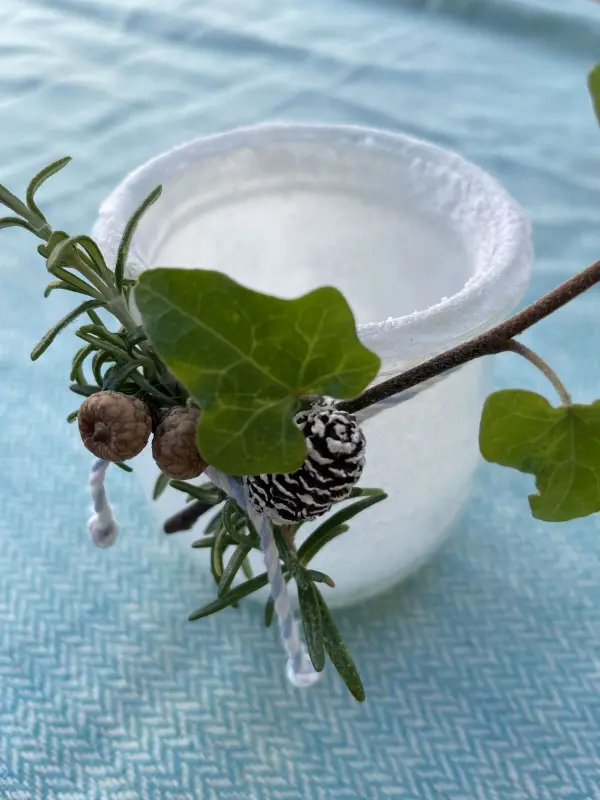 Mit einfachen Mitteln wie Salz zauberhafte Windlichter gestalten. Die Kristalle verleihen dem Glas einen frostigen Look. Dekoriere mit Naturmaterialien wie Efeu und Zweigen für ein stimmungsvolles Ambiente. Ideal für jede Jahreszeit. Ein Windlicht aus Glas, mit weißen gewachsenen Salzkristallen verziert, umgeben von einem grünen Blatt und kleinen Zapfen.