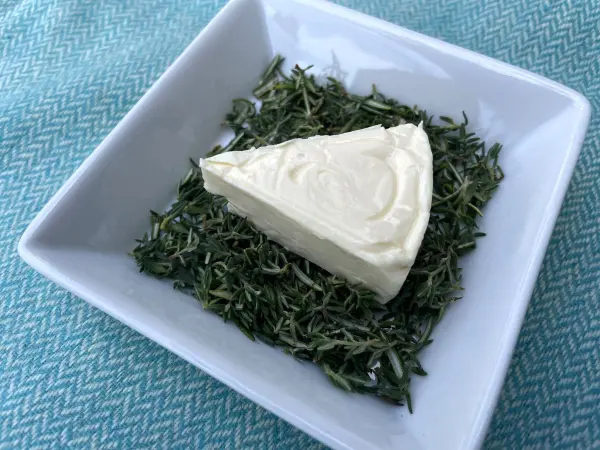 Kreiere mit Frischkäse-Tannenbäumen eine festliche Vorspeise. Das Bild zeigt, wie eine Frischkäse-Ecke in Kräutern gewendet wird. Ein Stück Frischkäse liegt auf einem Bett aus frischen Kräutern in einer weißen quadratischen Schale.