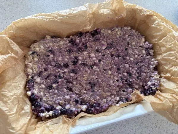 Die Haferflocken-Masse in eine kleine Auflaufform geben. Ich habe ein Backpapier benutzt, da ich so hinterher die Stücke besser schneiden kann. Haferflocken mit Heidelbeeren und Hüttenkäse in einer mit Backpapier ausgelegten Auflaufform auf heller Küchenarbeitsplatte.