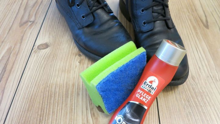 Ein K&uuml;chenschwamm liegt neben einer Flasche Schuhcreme und schwarzen Schuhen auf einem Holzfu&szlig;boden, bereit zum Eincremen.