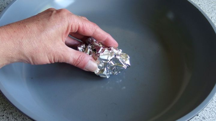 Eine Hand h&auml;lt ein St&uuml;ck Alufolie, das in einer grauen Pfanne mit Wasser verwendet wird, um die Pfanne zu reinigen.