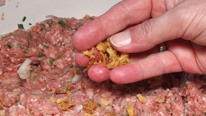 Eine Hand streut R&ouml;stzwiebeln &uuml;ber eine Mischung aus Hackfleisch, um Frikadellen zu w&uuml;rzen und geschmacklich zu verbessern.