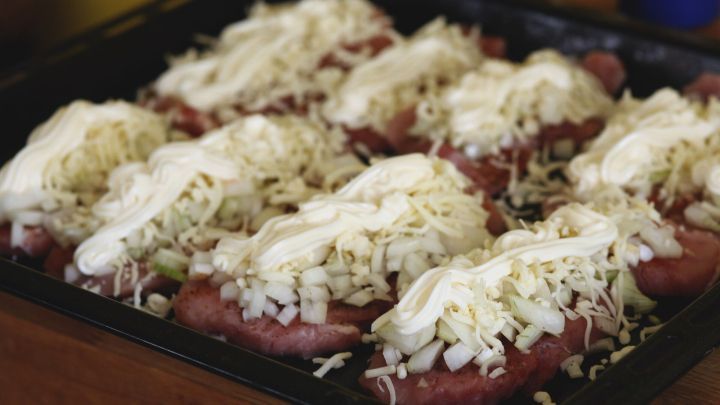 Zarte Schnitzel sind mit Zwiebeln, K&auml;se und Mayo belegt und auf einem Blech f&uuml;r die Zubereitung platziert.