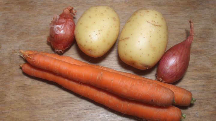 Ein Zwiebeltopf zeigt Karotten, Kartoffeln und Schalotten, die auf einer Holzoberfl&auml;che angeordnet sind, bereit zum Kochen.