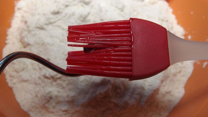 Ein roter Silikon-Pinsel kr&uuml;melt Mehl in einer orangefarbenen Sch&uuml;ssel, passend zum Thema M&uuml;rbeteig und K&uuml;chenutensilien.
