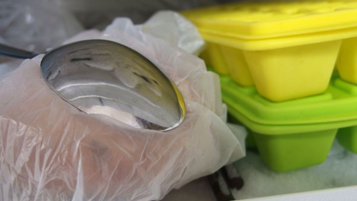 Eine Hand mit einem L&ouml;ffel, eingeh&uuml;llt in eine Plastikhandschuh, greift nach einem Gelbeisbeh&auml;lter im Gefrierfach.