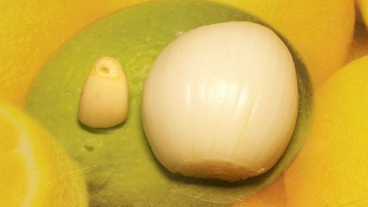 Eine wei&szlig;e Zwiebel und eine Knoblauchzehe liegen auf einer gr&uuml;nen Unterlage, umgeben von gelbem Hintergrund.