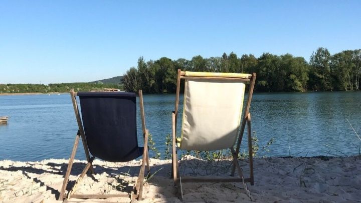 St&uuml;hle stehen am Sandstrand, w&auml;hrend die Sonne auf einen ruhigen See scheint, ideal f&uuml;r ein K&uuml;hles Bier am See.