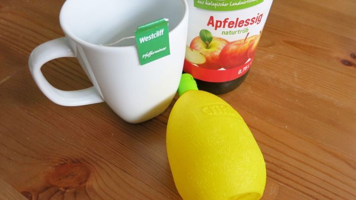 Eine Tasse mit Teebeutel steht neben einer Flasche Apfelessig und einer gelben Zitronenpressform auf einem Holzuntergrund.