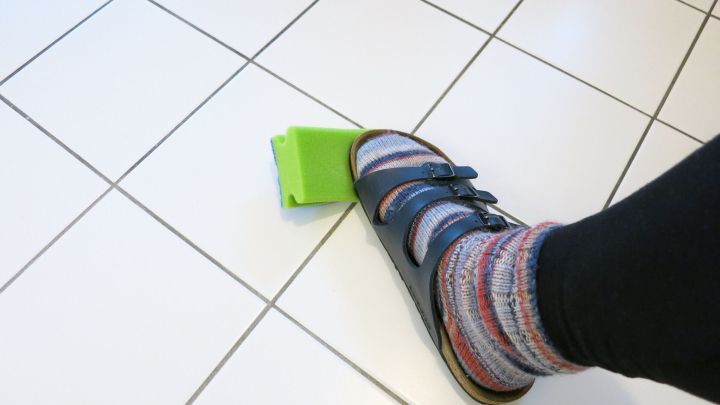 Ein Fu&szlig; mit einem bunten Socken dr&uuml;ckt auf einen gr&uuml;nen Wischmop auf einem glatten Boden, w&auml;hrend der Raum hell erleuchtet ist.