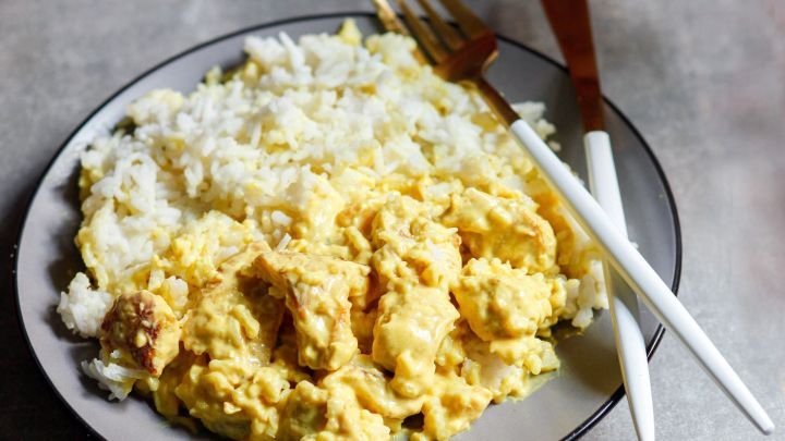 Ein Teller mit geschnetzeltem H&auml;hnchen in einer cremigen Sauce liegt neben dampfendem Reis auf einem grauen Tisch.