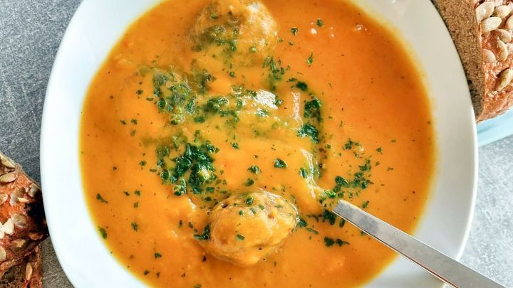 Eine cremige M&ouml;hrencremesuppe mit gr&uuml;ner Petersilie und Fleischb&auml;llchen in einer wei&szlig;en Sch&uuml;ssel auf einem grauen Tisch.