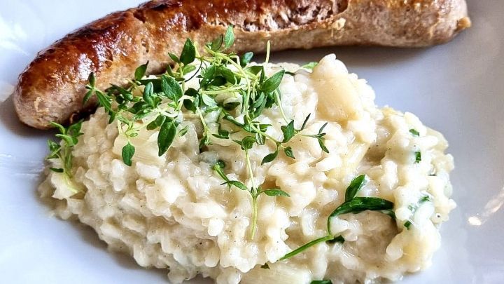 W&uuml;rzige Bratwurst liegt neben cremigem Risotto mit Kr&auml;utern garniert auf einem wei&szlig;en Teller, ready to enjoy.
