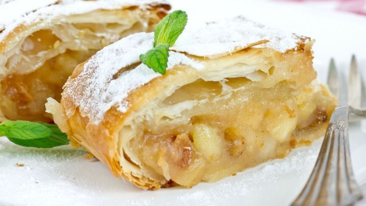 Zwei St&uuml;cke Apfelstrudel mit Puderzucker und Minze liegen auf einem wei&szlig;en Teller vor einem karierten Hintergrund.