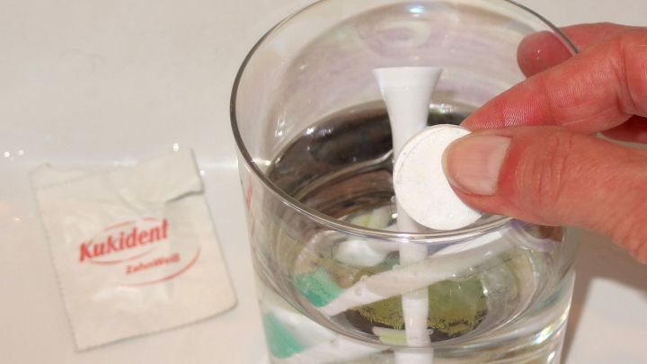 Eine Hand gibt eine Reinigungstablette in ein Glas Wasser, das einen Zahnb&uuml;rstenaufsatz mit Gebissreiniger enth&auml;lt.