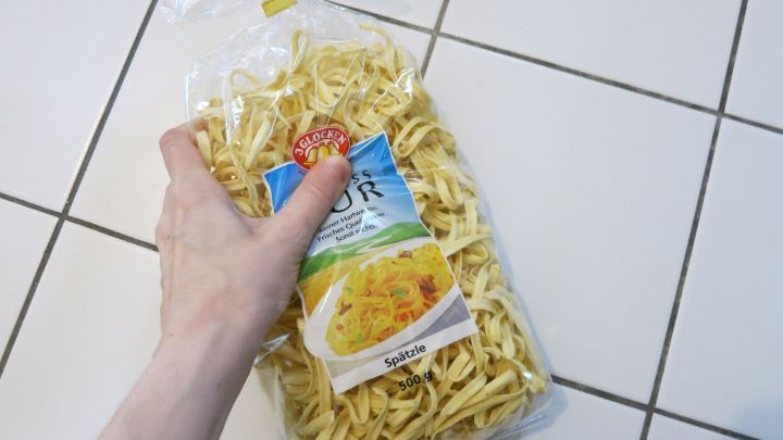 Eine Hand h&auml;lt eine T&uuml;te Sp&auml;tzle in der K&uuml;che, bereit zum Kochen und Verzehr. Die Fliesen bilden den Hintergrund.