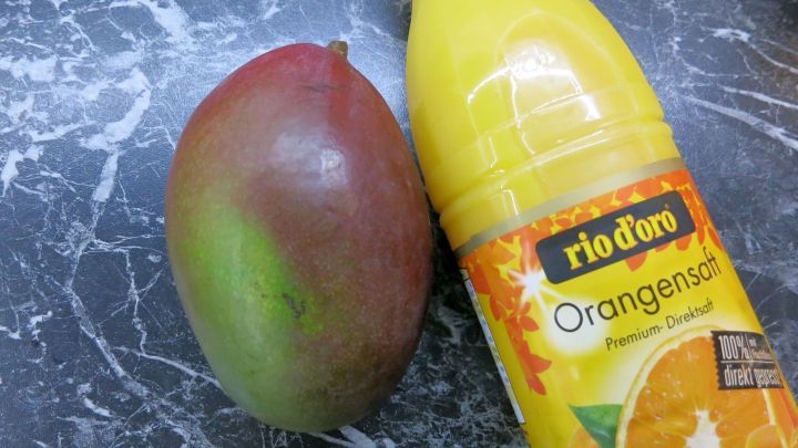 Eine reife Mango liegt neben einer gelben Flasche mit Orangensaft auf einer dunklen, marmorierten Oberfl&auml;che.