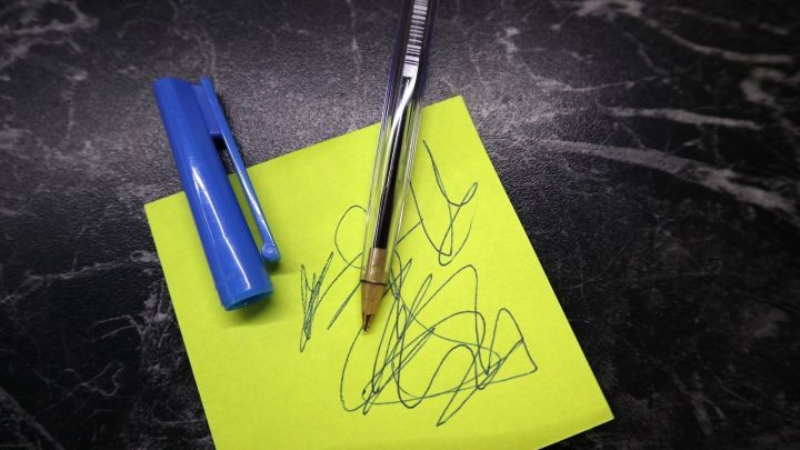 Ein Kugelschreiber liegt auf einem leuchtend gelben Haftnotizblock, umgeben von schwachem Licht. Kugelschreiberflecken sind sichtbar.