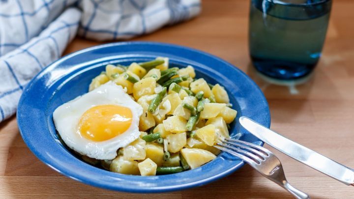 Ein Teller mit Kartoffel-Bohnen-Durcheinander steht auf einem Tisch, erg&auml;nzt durch ein Spiegelei und ein Glas Wasser.