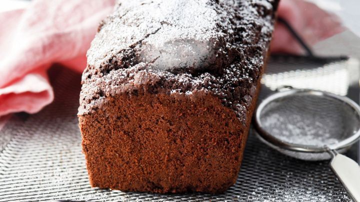 Ein saftiger Schokoladenkuchen liegt auf einem Kuchengitter, umgeben von einem rosa Tuch und einem Sieb.