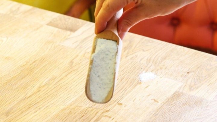 Eine Hand streut Waschpulver auf einen &Ouml;lfleck auf einem Holztisch. Der Hintergrund zeigt eine hellgelbe Wand und einen roten Stuhl.