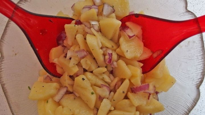 In einer Sch&uuml;ssel liegt ein Kartoffelsalat, der mit roten Zwiebeln und Gew&uuml;rzen vermischt wird, w&auml;hrend zwei rote L&ouml;ffel ihn umr&uuml;hren.