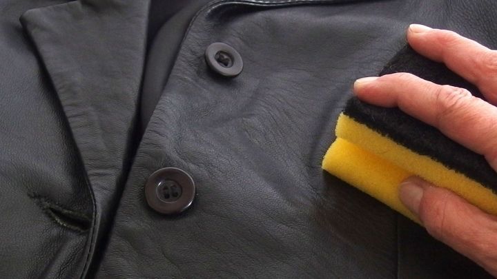 Eine Hand reinigt eine schwarze Lederjacke mit einem gelben Schwamm, um die Pflege der Jacke zu sichern.